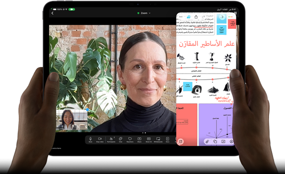 جهاز iPad Pro، الجزء الأمامي الخارجي، الوضع الأفقي، لون أسود فلكي، زوايا مدورة، إطار شاشة أسود، يدان تحملان الجهاز من الجانب الأيسر والأيمن، شاشة تعرض محادثة عبر فيس تايم تتم ترجمتها مباشرة، وتطبيق تعليمي يُظهر عرضاً تقديمياً بعنوان 'علم الأساطير المقارَن' مع نصوص ومخططات وملاحظات مكتوبة بخط اليد