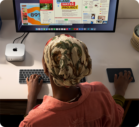 سيدة تطبع وهي جالسة إلى مكتب مع جهاز Mac mini، شاشة Studio Display متصلة، لوحة مفاتيح ماجيك، لوحة تعقب ماجيك، أغراض متنوعة