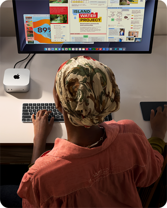 سيدة تطبع وهي جالسة إلى مكتب مع جهاز Mac mini، شاشة Studio Display متصلة، لوحة مفاتيح ماجيك، لوحة تعقب ماجيك، أغراض متنوعة