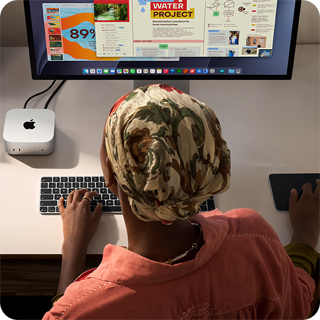 سيدة تطبع وهي جالسة إلى مكتب مع جهاز Mac mini، شاشة Studio Display متصلة، لوحة مفاتيح ماجيك، لوحة تعقب ماجيك، أغراض متنوعة