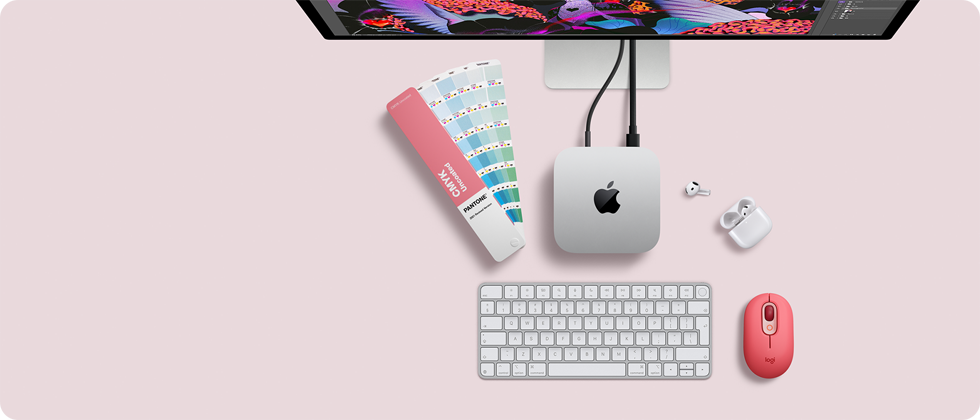 Mac mini, top, silver aluminum, rounded corners, glossy black Apple logo centered, connected by cord, Studio Display partially visible on top, AirPod 4, open, Pantone color swatches, and numeric keyboard and mouse