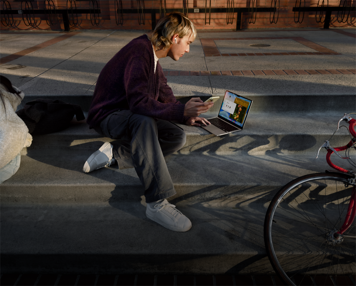 Person sitting on concrete steps outdoors, wearing an AirPod, holding an iPhone, and navigating on the trackpad of MacBook opened on the ground in front of them, red bicycle lying on floor nearby, another person sitting nearby