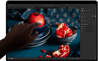 جهاز MacBook Pro مع شاشة PROLED XDR يعرض صورة ثابتة لثمار الرمان وقطع خزفية ومنسوجات زرقاء، مع يد تمتد نحو الشاشة للتفاعل مع العمل الفني